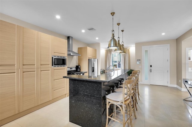 kitchen with a kitchen island with sink, light brown cabinets, wall chimney exhaust hood, stainless steel appliances, and decorative light fixtures