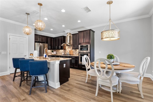 kitchen featuring a kitchen island with sink, visible vents, hanging light fixtures, appliances with stainless steel finishes, and wall chimney exhaust hood