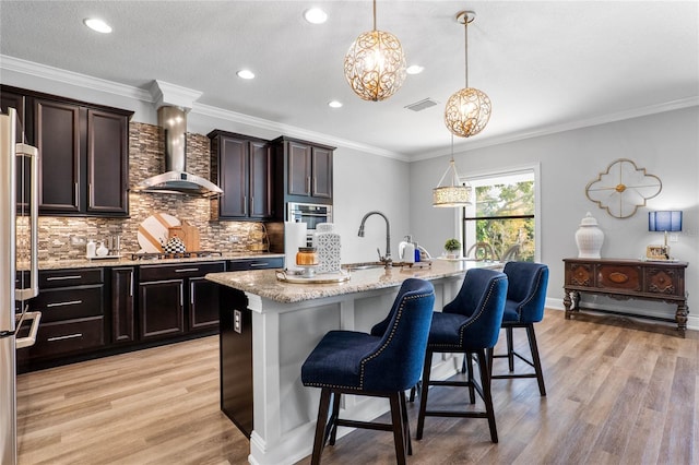 kitchen with wall chimney exhaust hood, hanging light fixtures, a sink, and a center island with sink