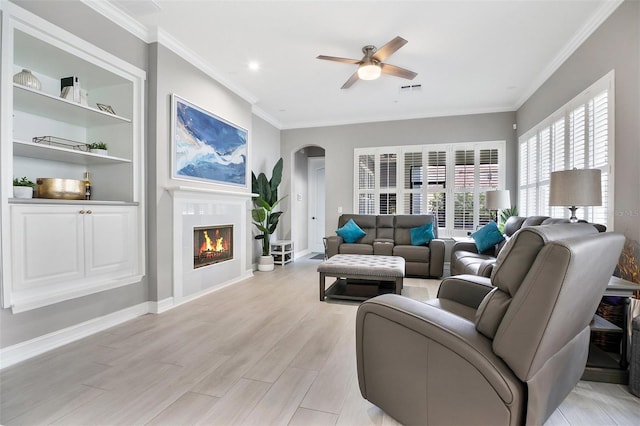 living room with crown molding, built in shelves, light wood-type flooring, and ceiling fan