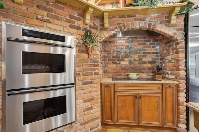 kitchen with black electric cooktop, double oven, light stone counters, and brick wall