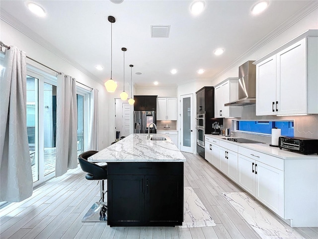 kitchen featuring wall chimney exhaust hood, hanging light fixtures, a center island with sink, white cabinets, and appliances with stainless steel finishes