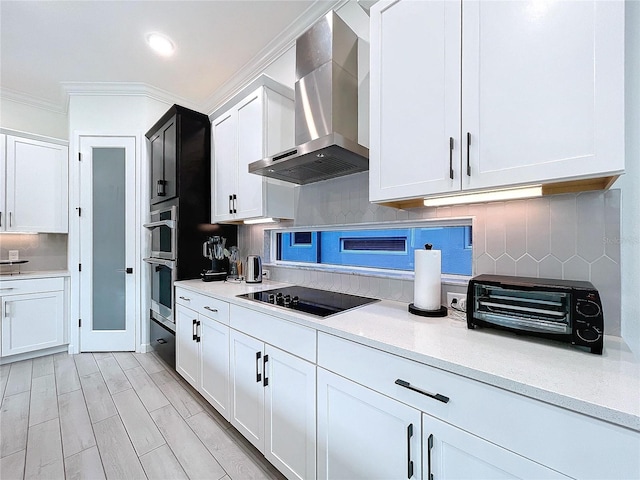 kitchen featuring wall chimney range hood, white cabinets, decorative backsplash, crown molding, and black electric stovetop