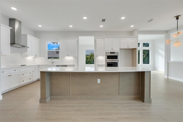 kitchen featuring a spacious island, wall chimney exhaust hood, stainless steel appliances, pendant lighting, and white cabinets