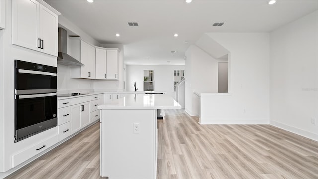 kitchen featuring wall chimney range hood, a kitchen island, white cabinetry, light wood-type flooring, and double oven