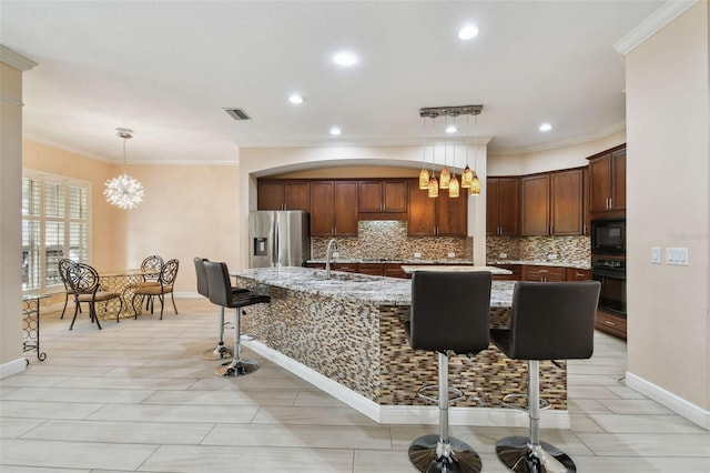 kitchen with decorative light fixtures, a kitchen breakfast bar, an island with sink, light stone countertops, and black appliances