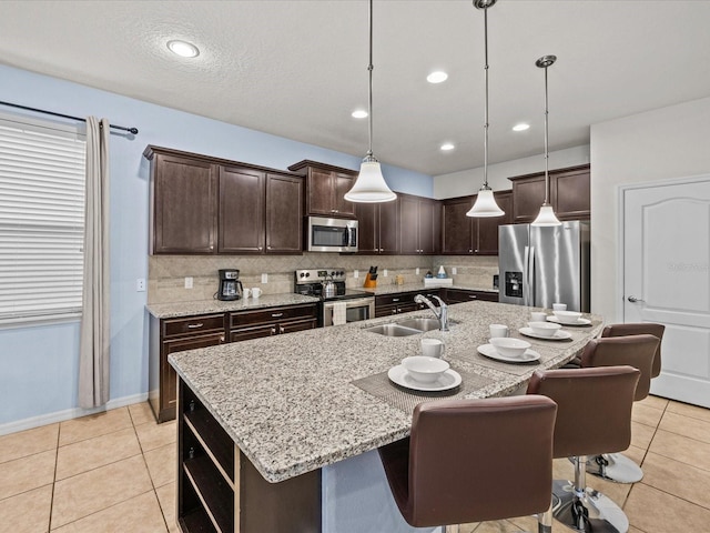 kitchen with an island with sink, dark brown cabinets, stainless steel appliances, sink, and pendant lighting