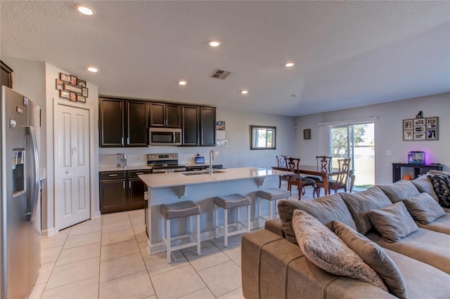 kitchen with light tile patterned floors, an island with sink, sink, dark brown cabinetry, and stainless steel appliances