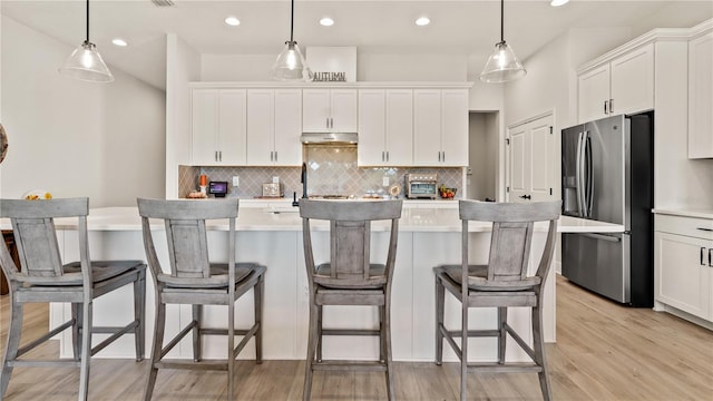 kitchen featuring white cabinets, light countertops, under cabinet range hood, stainless steel fridge, and backsplash