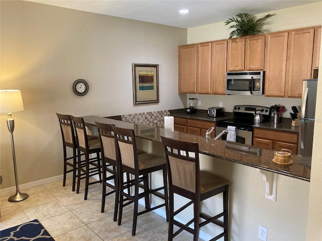 kitchen with appliances with stainless steel finishes, a kitchen bar, kitchen peninsula, dark stone counters, and light tile patterned floors