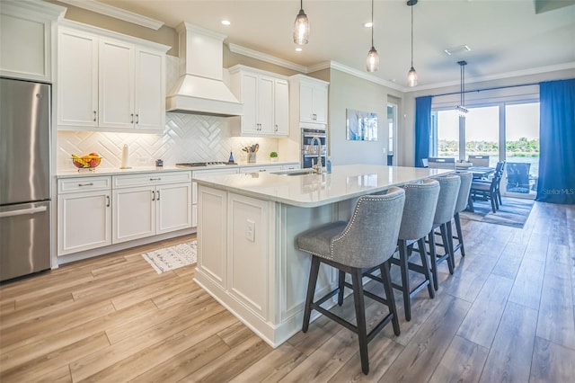 kitchen featuring a kitchen island with sink, premium range hood, stainless steel appliances, and white cabinetry