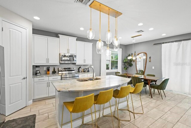 kitchen featuring hanging light fixtures, white cabinetry, stainless steel appliances, light stone counters, and a kitchen island with sink