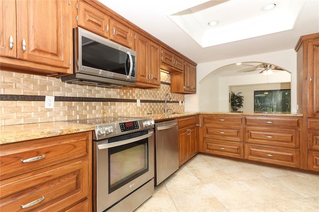 kitchen with appliances with stainless steel finishes, light stone counters, a tray ceiling, ceiling fan, and sink