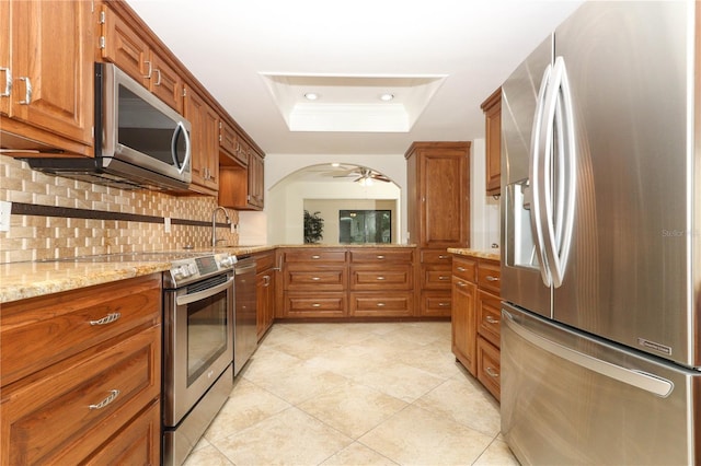 kitchen featuring ceiling fan, sink, stainless steel appliances, light stone counters, and a tray ceiling