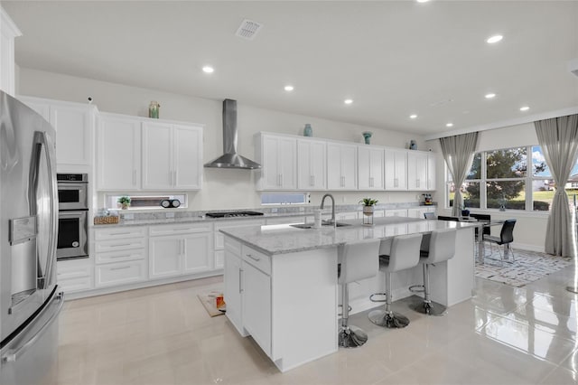 kitchen with sink, wall chimney exhaust hood, stainless steel appliances, a kitchen island with sink, and white cabinets