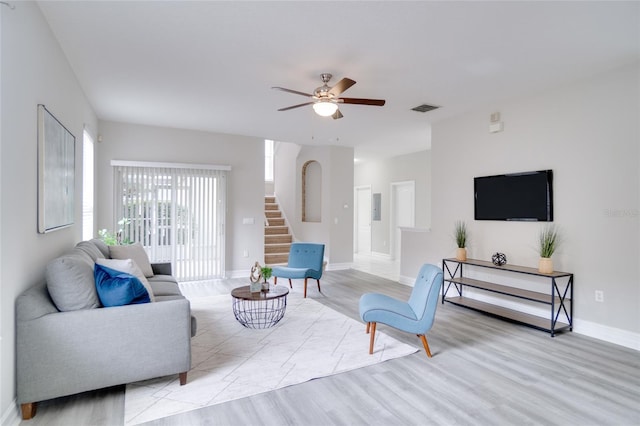 living room with ceiling fan, light wood-style flooring, baseboards, visible vents, and stairway
