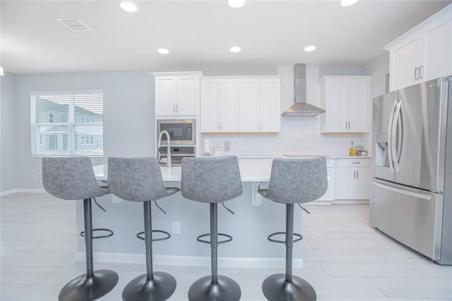 kitchen featuring wall chimney range hood, white cabinets, a center island with sink, appliances with stainless steel finishes, and a kitchen breakfast bar