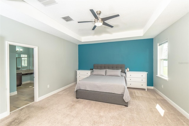 bedroom featuring ensuite bathroom, light colored carpet, a raised ceiling, and ceiling fan