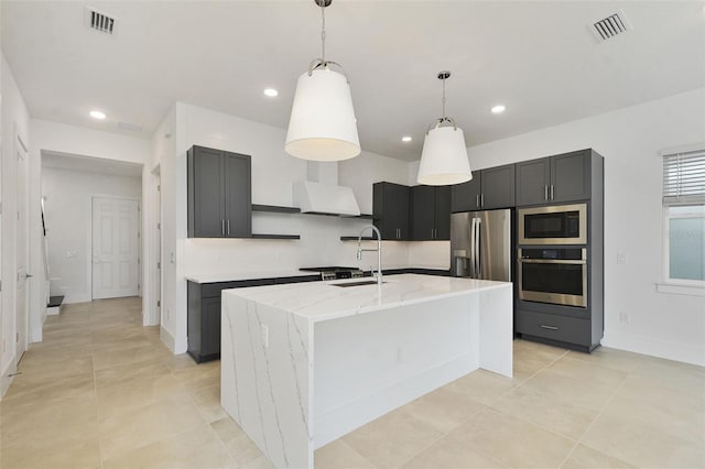 kitchen featuring appliances with stainless steel finishes, sink, a kitchen island with sink, light stone counters, and custom range hood