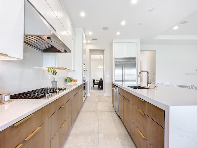 kitchen featuring a large island with sink, wall chimney range hood, sink, appliances with stainless steel finishes, and white cabinets