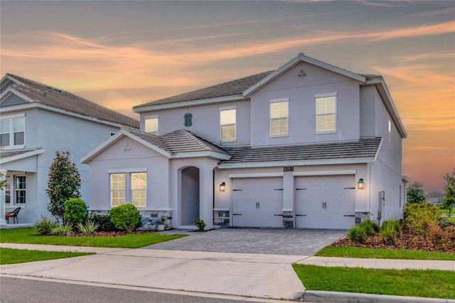 traditional-style home featuring a garage, a tile roof, stone siding, decorative driveway, and stucco siding