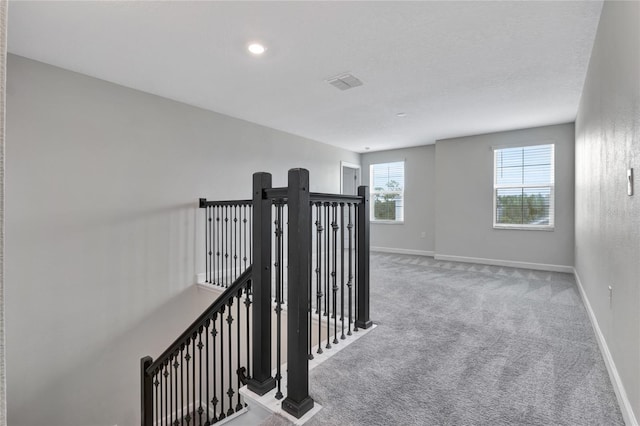 hallway with an upstairs landing, baseboards, visible vents, and light colored carpet