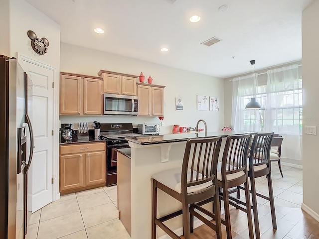 kitchen with light brown cabinets, appliances with stainless steel finishes, a kitchen breakfast bar, a kitchen island with sink, and pendant lighting