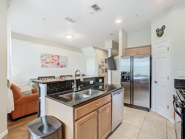 kitchen with appliances with stainless steel finishes, light tile patterned flooring, sink, an island with sink, and dark stone countertops