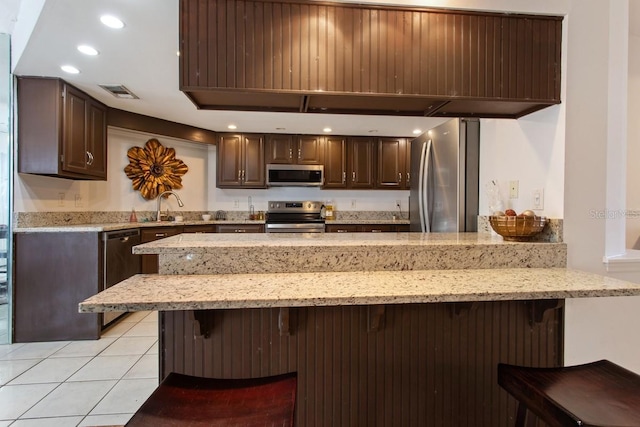 kitchen with light tile patterned flooring, stainless steel appliances, dark brown cabinets, and a kitchen bar