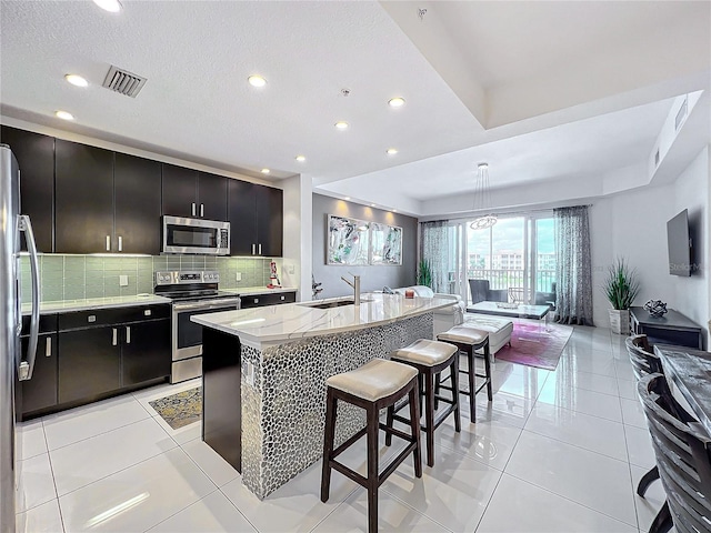 kitchen featuring backsplash, a kitchen island with sink, sink, appliances with stainless steel finishes, and light tile patterned flooring