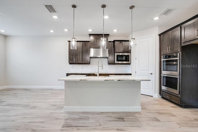 kitchen featuring double oven, built in microwave, hanging light fixtures, dark brown cabinetry, and a center island with sink