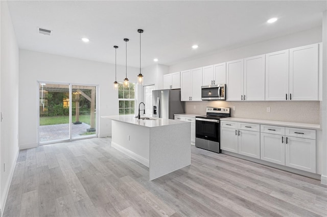 kitchen with a kitchen island with sink, stainless steel appliances, pendant lighting, light wood-type flooring, and white cabinets