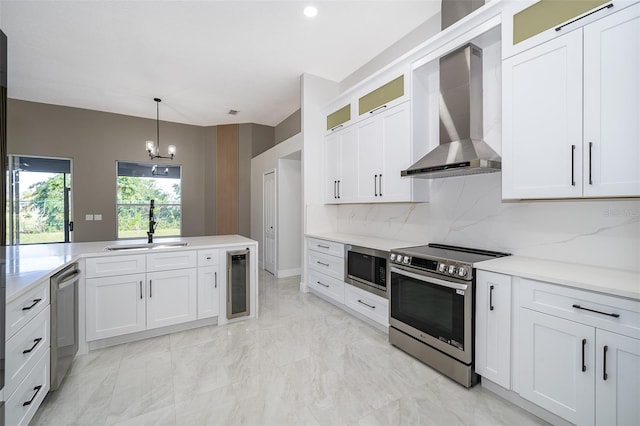 kitchen featuring wall chimney range hood, sink, white cabinetry, stainless steel appliances, and decorative light fixtures