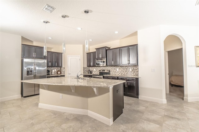 kitchen with an island with sink, hanging light fixtures, stainless steel appliances, light stone countertops, and tasteful backsplash