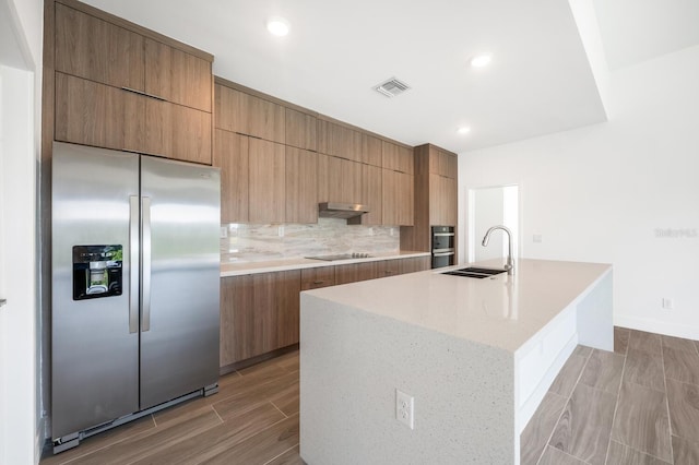 kitchen with sink, stainless steel fridge, black electric stovetop, a kitchen island with sink, and decorative backsplash