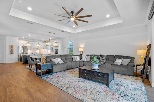 living room featuring ceiling fan with notable chandelier, a tray ceiling, and light wood-type flooring