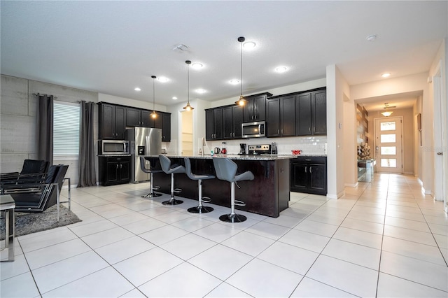 kitchen with stainless steel appliances, a center island with sink, light stone countertops, a breakfast bar area, and hanging light fixtures