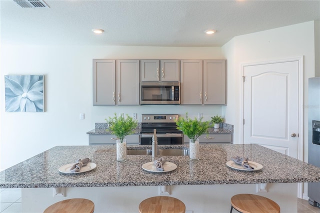 kitchen with gray cabinetry, appliances with stainless steel finishes, stone countertops, and a kitchen island with sink