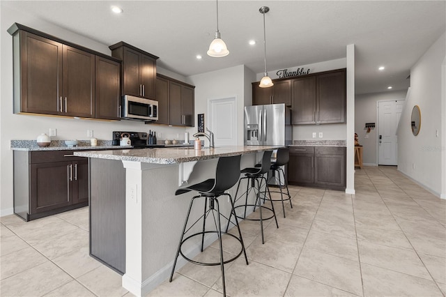 kitchen featuring light stone countertops, stainless steel appliances, dark brown cabinetry, pendant lighting, and a center island with sink