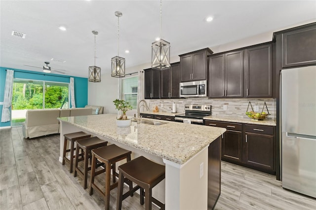 kitchen featuring hanging light fixtures, appliances with stainless steel finishes, a kitchen island with sink, light wood-type flooring, and sink