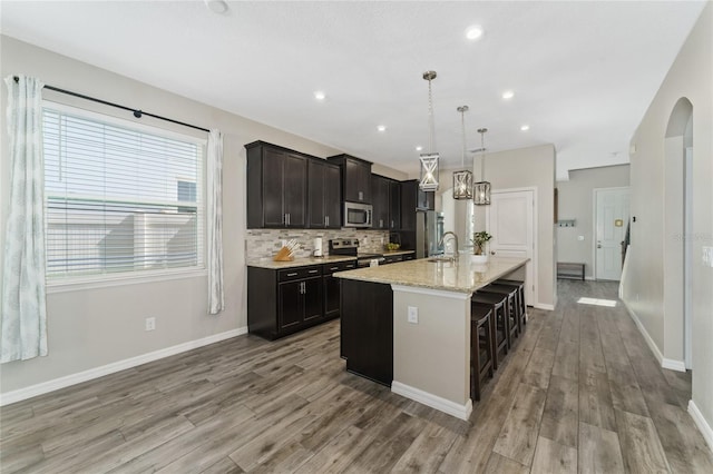 kitchen featuring a kitchen island with sink, light hardwood / wood-style flooring, hanging light fixtures, stainless steel appliances, and light stone countertops