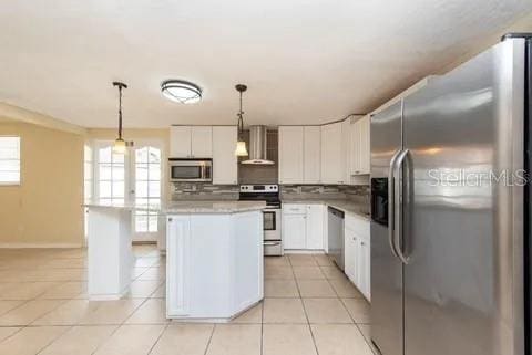 kitchen with wall chimney range hood, decorative backsplash, white cabinets, hanging light fixtures, and stainless steel appliances