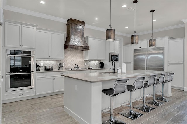 kitchen featuring built in appliances, custom exhaust hood, white cabinetry, and a kitchen island with sink