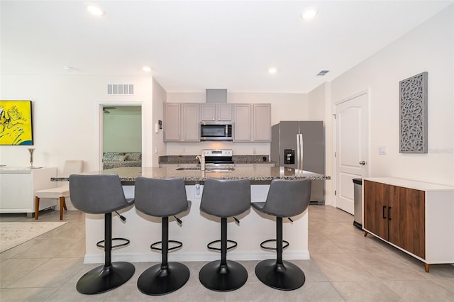 kitchen featuring light tile patterned floors, appliances with stainless steel finishes, a kitchen island with sink, light stone countertops, and gray cabinets