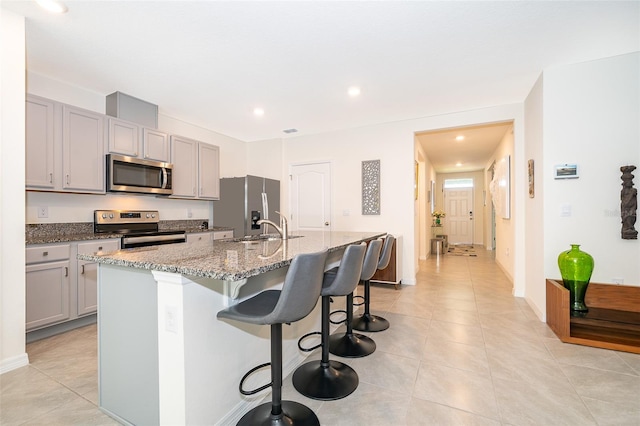 kitchen with gray cabinetry, stone counters, appliances with stainless steel finishes, a breakfast bar area, and a center island with sink