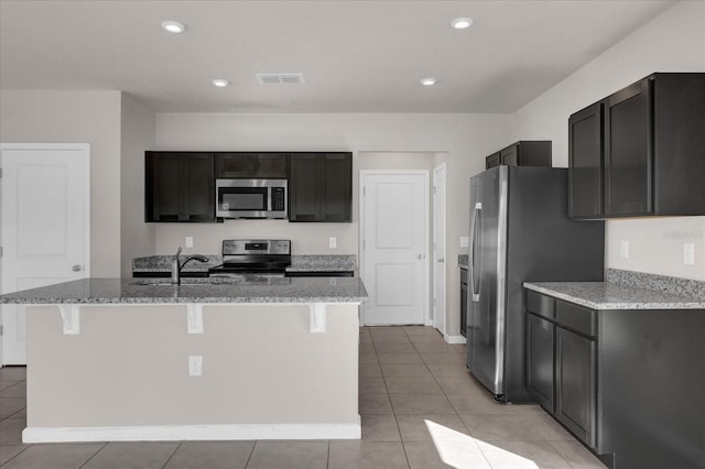 kitchen with light stone counters, light tile patterned floors, an island with sink, and appliances with stainless steel finishes