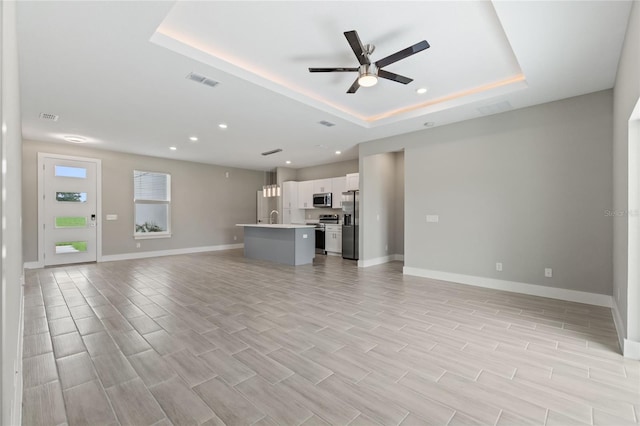 unfurnished living room featuring light hardwood / wood-style floors, ceiling fan, sink, and a tray ceiling