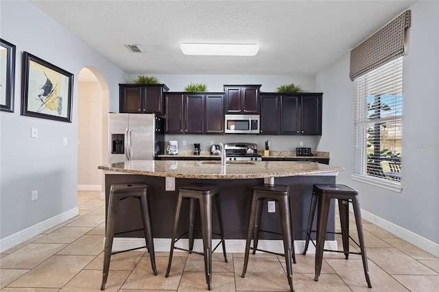 kitchen featuring stainless steel appliances, a center island with sink, a textured ceiling, a breakfast bar, and dark brown cabinets