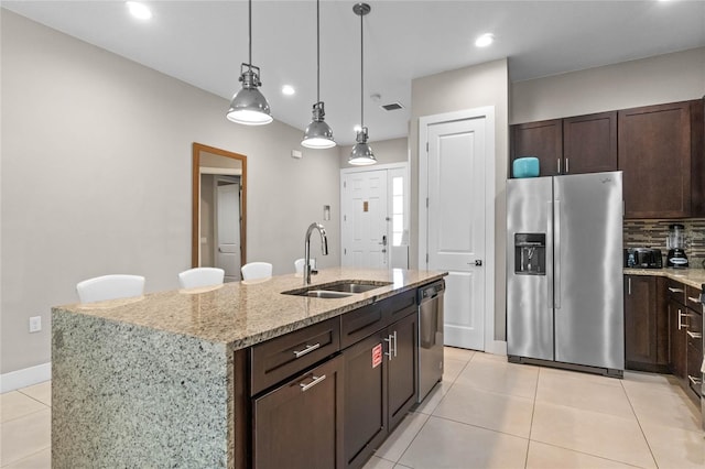 kitchen featuring appliances with stainless steel finishes, light stone counters, dark brown cabinets, a kitchen island with sink, and sink