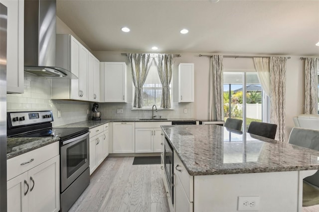 kitchen featuring a kitchen island, electric stove, wall chimney range hood, white cabinets, and a breakfast bar area